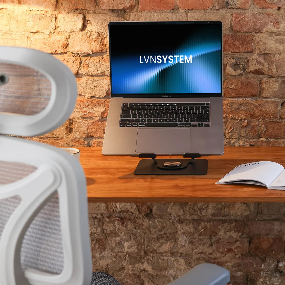 Laptop na podstawce Varia Flex na drewnianym biurku przed ścianą z cegły